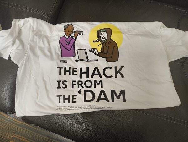 A white T-shirt printed with a cartoon of a man in a purple sweater looking through a telescope at a hooded hacker typing on a laptop, above the text THE HACK IS FROM THE 'DAM